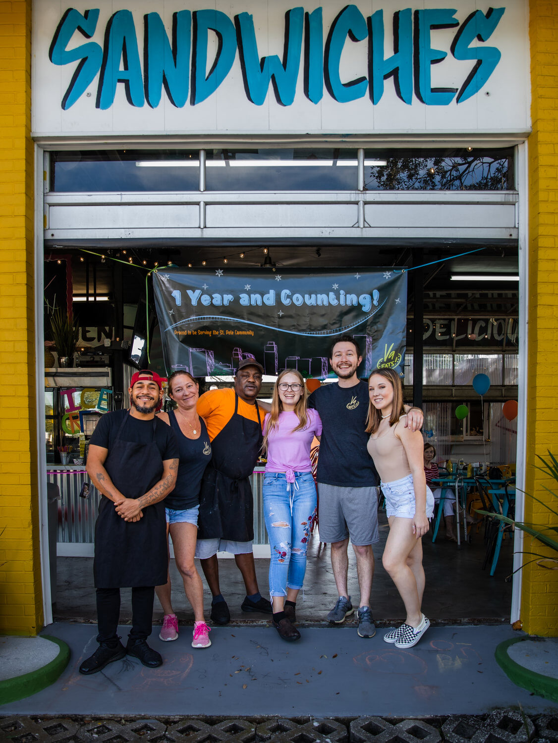 The V9 EATS crew standing at the SANDWICHES garage door under a '1 Year and Counting' banner